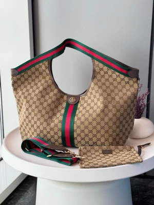 Gucci beige monogram handväska - Säljer en beige handväska från Gucci med klassiskt GG-monogram, gröna och röda ränder samt guldfärgade detaljer. Väskan har bred axelrem och matchande liten clutch. Tillverkad i canvas med läderdetaljer. Perfekt för dig som gillar lyxig och tidlös design.