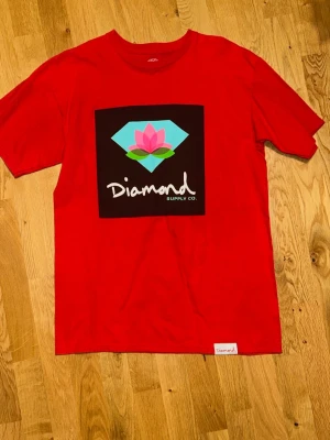 Röd Diamond Supply Co t-shirt - Snygg röd t-shirt från Diamond Supply Co med stort tryck framtill av en blå diamant och rosa blomma. Klassisk passform och korta ärmar. Tillverkad i mjuk bomull, perfekt för en avslappnad streetwear-look.