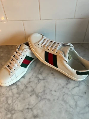 Gucci vita sneakers med röd/grön rand - Säljer ett par klassiska vita sneakers från Gucci med de ikoniska gröna och röda ränderna på sidan. Skorna har vita snören, rund tå och är tillverkade i slätt läder. Perfekta för dig som vill ha en clean och stilren look med lyxig känsla.