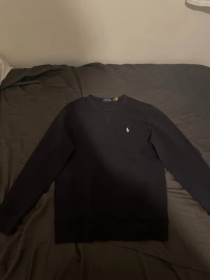Svart sweatshirt från Polo Ralph Lauren - Klassisk svart sweatshirt från Polo Ralph Lauren med rund halsringning och den ikoniska vita logon broderad på bröstet. Tröjan har långa ärmar och ribbade muddar vid ärmslut och nederkant. Perfekt för en stilren och avslappnad look.