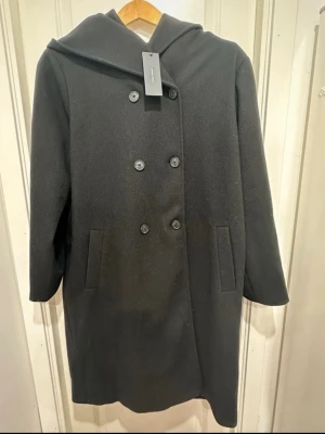 Svart dubbelknäppt kappa Vero Moda - Stilren svart kappa från Vero Moda i storlek XL. Kappan har dubbelknäppning med sex knappar, två sidofickor och en klassisk huva. Tillverkad i ett mjukt ull-liknande material som håller dig varm under kyliga dagar. Perfekt för dig som gillar minimalistisk och tidlös design.