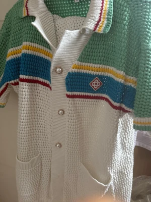 Casablanca sommar kläder tröja o shorts stickad - Säljer ett stickat set med kortärmad body och shorts i vitt, grönt, blått, gult och rött. Setet har pärlemorknappar, krage, fickor och ett broderat märke på bröstet. Materialet är grovstickad bomull och plagget har sportiga ränder för en retrovibe.