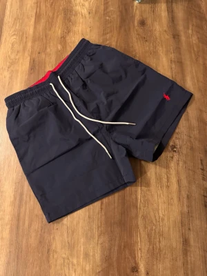 Marinblå badshorts från Polo Ralph Lauren - Snygga marinblå badshorts från Polo Ralph Lauren med röd logga broderad på benet. Shortsen har vit dragsko i midjan, sidofickor och en bakficka med knapp. Insidan är fodrad med rött meshfoder. Perfekta för stranden eller poolen.