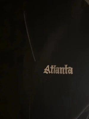 Svart hoodie  - Säljer en svart hoodie med Atlanta-tryck i vitt framtill och stort tryck på ryggen där det står 'Property of Atlanta Athletic Dept'. Klassisk modell med huva och känguruficka. Perfekt för dig som gillar streetwear och sportig stil.