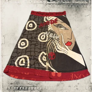 Färgstark kjol med grafiskt tryck - Unik kjol med grafiskt ansiktstryck, textdetaljer och röda kontrastkanter. Kjolen har en A-linjeform och elastisk midja. Färgerna går i brunt, rött, beige och svart med inslag av vitt. Perfekt för dig som gillar statement-plagg och konstnärlig stil.