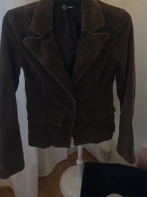 Brun manchesterkavaj från ONLY - Snygg brun kavaj i manchestertyg från ONLY med klassisk krage och två knappar framtill. Kavajen har fickor med lock och markerad midja för en schysst siluett. Perfekt för dig som vill ha en trendig och tidlös look.