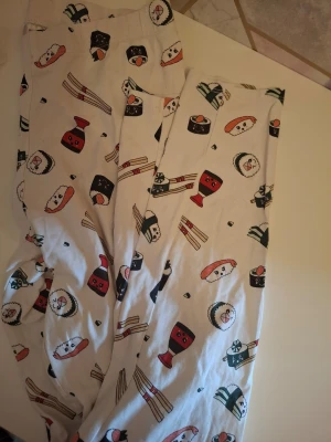 Sushimönstrade pyjamasbyxor H&M Divided - Roliga pyjamasbyxor från H&M Divided med ett färgglatt sushimönster. Byxorna är vita med tryck av olika sushibitar, sojaflaskor och ätpinnar. Mjukt och bekvämt material, perfekt för mysiga hemmakvällar. Resår i midjan för extra komfort.