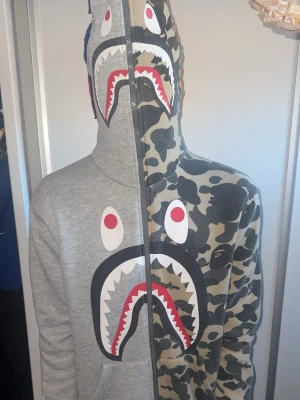 BAPE Shark hoodie camo/grå - Unik BAPE Shark hoodie med dragkedja hela vägen upp i huvan. Ena sidan är grå och andra sidan har klassiskt camomönster. Stort hajtryck med ögon och tänder framtill. Tillverkad i mjuk bomull, har känguruficka och ribbade muddar.