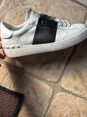 Vit och svart sneaker från Valentino - Snygg sneaker i vitt läder med svart detalj på sidan och nitar vid hälen. Klassisk låg modell med vita skosnören och tjock vit sula. Perfekt för dig som gillar stilrena och exklusiva sneakers.