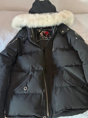Svart dunjacka med päls från Moose Knuckles - Svart herr dunjacka från Moose Knuckles med stor huva och vit pälskant. I princip aldrig använd, ny kostade 12000 är alltså ÄKTA, kvitto finns och följer med vid köp