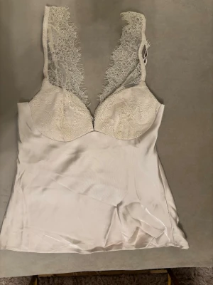 Champagne färgad spetsdetaljerad linnetopp - Snygg vit linnetopp med spetsdetaljer vid axelbanden och kuporna. Tillverkad i ett glansigt, silkesliknande material som ger en lyxig känsla. Perfekt att styla med jeans eller kjol för en trendig look.