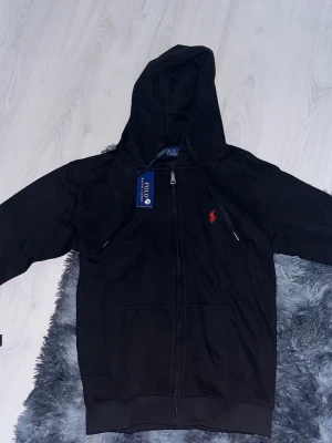 Svart hoodie från Polo Ralph Lauren - Svart hoodie med dragkedja från Polo Ralph Lauren. Klassisk design med huva och fickor framtill. Liten röd logga broderad på bröstet. Tillverkad i mjukt bomullsmaterial som är skönt att bära. Perfekt för en avslappnad och stilren look.