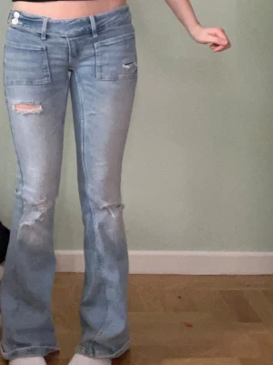 Gina jeans - Säljer ett par ljusblå bootcut jeans med låg midja och slitna detaljer på benen. Jeansen har stora framfickor och dubbla knappar i linningen. Perfekta för en avslappnad och trendig look.