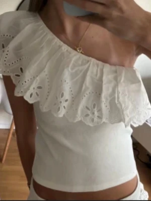Vit oneshoulder topp med volang - Säljer en vit oneshoulder topp med stor broderad volangkant och hålmönster. Toppen är croppad och har en tight passform, perfekt till jeans eller kjol. Materialet känns som bomull och volangen ger en romantisk vibe.