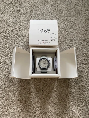 Omega x Swatch Moonswatch 1965 - Säljer en Omega x Swatch Moonswatch Mission To The Moon 1965 Special Edition från Bioceramic Collection. Klockan har en grå boett, svart tachymeter-ring och vit urtavla med svarta detaljer. Snygg och modern design med klassiska Speedmaster-vibbar. Kommer i originalbox med tillbehör.