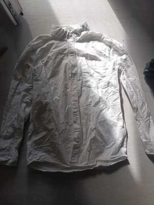 Vit långärmad skjorta med ficka - Klassisk vit skjorta med lång ärm och bröstficka. Skjortan har knappar framtill och en snygg krage. Perfekt till jeans eller chinos för en clean look. Tillverkad i bomull för skön känsla och andningsförmåga.