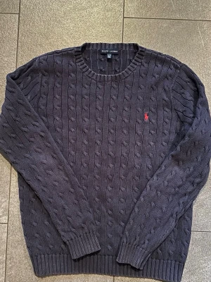 Mörkblå kabelstickad tröja Ralph Lauren - Kabelstickad mörkblå tröja från Ralph Lauren med rund halsringning och klassisk röd broderad logga på bröstet. Tröjan har ribbade muddar vid ärmslut och nederkant, perfekt för dig som gillar stilren och tidlös design.