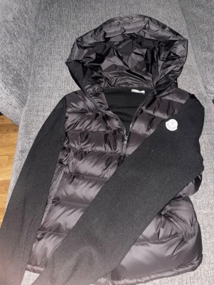 Svart Moncler cardiganjacka med huva - Snygg svart cardiganjacka från Moncler med vadderad kropp och stickade ärmar. Jackan har en stor huva och dragkedja framtill samt Moncler-logga på bröstet. Perfekt för lager på lager och streetstyle. Materialmix av dun och stickat ger både värme och stil. Äkta moncler cardigan säljs för 1000kr. OBS PRISET ÄR INTE FAST!!!