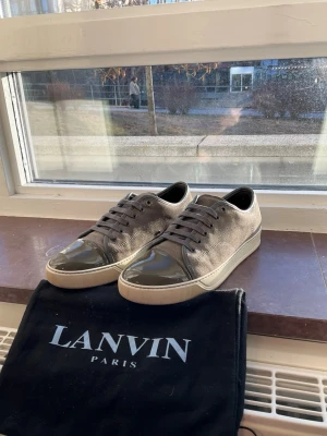 Grå gröna sneakers från Lanvin - Snygga grå sneakers från Lanvin med glansig tå i mörkgrått lack och ovandel i mocka. Skorna har vita sulor och matchande grå skosnören. Perfekta för dig som vill ha en stilren och exklusiv look. Väldigt fint skick och kommer med dustnag, storlek 42.