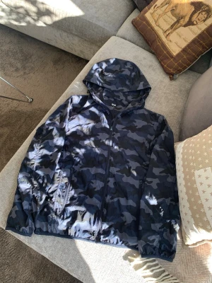 Mörkblå camo vindjacka med huva från Uniqlo - Snygg mörkblå vindjacka med camouflagemönster och huva. Jackan har dragkedja framtill, två sidofickor med dragkedja och elastiska muddar vid ärmarna. Perfekt för blåsiga dagar och enkel att packa ner i väskan.