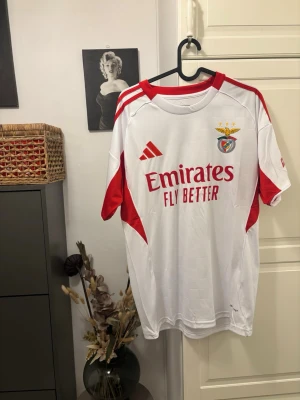 Vit Benfica matchtröja Adidas - Snygg vit Benfica matchtröja från Adidas med röda detaljer och klubbmärke på bröstet. Tryck med Emirates Fly Better på framsidan och Sagres på ryggen. Perfekt för fotbollsfans som vill visa sitt stöd för laget.