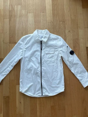 Vit overshirt från C.P. Company - Stilren vit overshirt från C.P. Company med svart dragkedja framtill, bröstficka och den ikoniska linsen på ärmen. Skjortjackan har lång ärm och klassisk krage, perfekt för lager på lager. Tillverkad i bomull för en skön känsla.