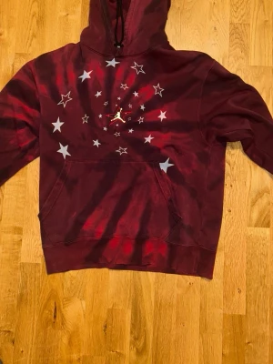 Jordan Vinröd tie-dye hoodie med stjärnor - Säljer en vinröd och svart tie-dye hoodie från Jordan med huva och känguruficka. Framsidan har ett coolt stjärnmönster i vitt och silver runt det klassiska Jumpman-loggan. Perfekt för dig som vill sticka ut med streetwear vibes.