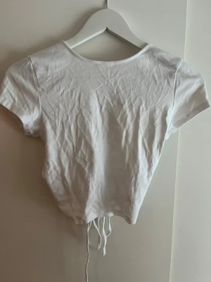 Vit croppad topp med snörning - Trendig vit croppad topp med korta ärmar och snygg snörning i ryggen som ger en öppen rygg-effekt. Toppen har en tight passform och är gjord i mjuk bomull, perfekt för att styla med jeans eller kjol.