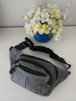 Grå midjeväska med flera fack - Snygg grå midjeväska i textil med flera dragkedjeförsedda fack och justerbart svart band. Perfekt för att bära det viktigaste och enkel att ha runt midjan eller över axeln. Stilren och praktisk design utan synligt märke. Helt ny 