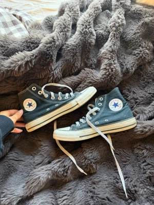 Svarta Converse Chuck Taylor Highs - Klassiska svarta Converse Chuck Taylor All Star high tops med vit sula och tå, vita skosnören och metallöljetter. Ikonisk logga på sidan och retrovibe som alltid funkar till streetwear. Canvasmaterial och tidlös design.