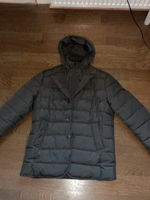 Svart dunjacka med huva från John Henric - Snygg mörkgrön dunjacka från John Henric med quiltad design och tre knappar framtill. Jackan har en avtagbar huva och klassisk krage, vilket ger en modern och stilren look. Tillverkad i lätt och värmande material, perfekt för kyliga dagar. Riktigt schysst samt mogen stil nästan lite oldmoney vibe. Riktigt bra passform också på denna. Jackan kosta från början 2000 och är i gott skick.