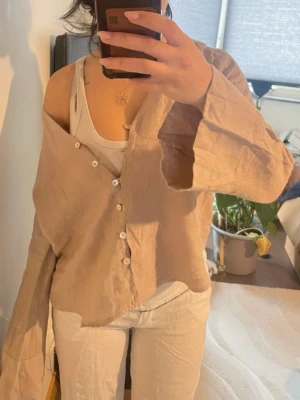 Beige linneskjorta från Zara - Säljer en beige skjorta från Zara i luftigt linne. Skjortan har klassisk krage, långa ärmar och vita knappar framtill. Passformen är loose och materialet känns svalt och skönt, perfekt för varmare dagar.