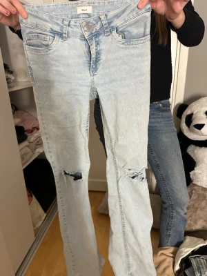 Ljusblå slitna bootcut jeans MLW - Säljer ett par ljusblå bootcut jeans från Nelly  med slitningar och hål på knäna. Jeansen har fem fickor, rosa sömmar på bakfickorna och klassisk knappgylf. Perfekta för en avslappnad och trendig look.