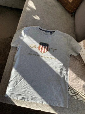 Grå GANT t-shirt med tryck - Snygg grå t-shirt från GANT med stort tryck framtill i guld och rött, samt ett sköldmärke med texten '1949' och 'American Sportswear'. Klassisk rund hals och korta ärmar. Perfekt för en avslappnad och sportig stil. Storlek 158/164, 13-14 år.