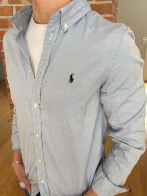 Ljusblå skjorta från Ralph Lauren - Klassisk ljusblå skjorta från Ralph Lauren med button-down krage och broderad logga på bröstet. Skjortan har långa ärmar, vita knappar och är tillverkad i mjuk bomull. Perfekt för en stilren och avslappnad look.