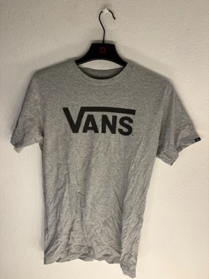 Grå T-shirt från Vans - Snygg grå T-shirt från Vans med klassisk svart logga tryckt på bröstet. T-shirten har rund halsringning och korta ärmar. Perfekt för en avslappnad streetwear-look och enkel att matcha med jeans eller shorts.