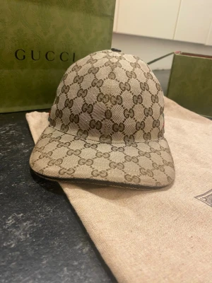 Gucci keps beige  - Säljer en beige keps från Gucci med det klassiska GG-monogrammönstret över hela kepsen. Kepsen har böjd skärm och detaljer i grönt och rött på sidan. Tillverkad i canvasmaterial med exklusiv känsla och ikonisk design.