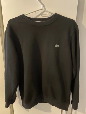 Svart sweatshirt från Lacoste - Klassisk svart sweatshirt från Lacoste med rund halsringning och den ikoniska gröna krokodilloggan broderad på bröstet. Tröjan har ribbade muddar vid ärmslut och nederkant. Perfekt för en clean och stilren look.