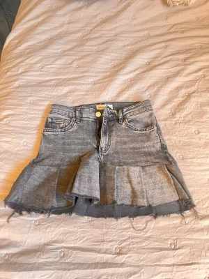 Grå jeanskjol med volangkant - Säljer en grå jeanskjol med volangkant och råa kanter. Kjolen har klassiska jeansdetaljer som fickor fram och bak, samt knapp och dragkedja i midjan. 