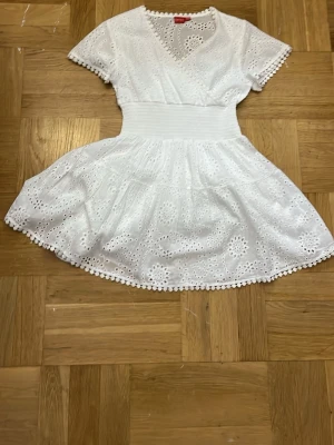 Vit kort klänning med spetsdetaljer - Söt vit kortklänning med v-ringning och broderade spetsdetaljer. Klänningen har korta ärmar, markerad midja och volangkant nertill. Använd 1 gång