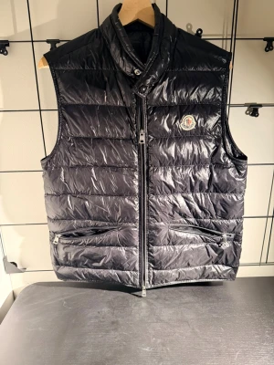Moncler Gui väst - Snygg svart dunväst från Moncler med glansig finish och klassisk quiltad design. Köpt på Kicks N Stuff. Utrustad med hög krage, två dragkedjefickor och ikonisk logga på bröstet. Perfekt för lager-på-lager och en stilren streetwear-look.  ✔ 100% äkta ✔ Köpt på Kicks N Stuff ✔ Lätt och varm ✔ Tidlös design