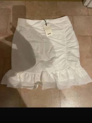 Vit volangkjol från Bik Bok - Snygg vit kjol från Bik Bok med volangkant nedtill och lätt rynkade detaljer längs sidorna. Kjolen har en kort passform och är perfekt för dig som gillar en fräsch och trendig look. Tillverkad i ett mjukt material som känns bekvämt hela dagen.