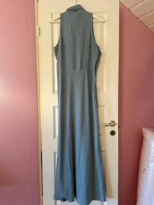 Ljusblå långklänning med knyt - Elegant ljusblå långklänning med hög hals och knyt i nacken. Klänningen är ärmlös och har en mjuk, flytande passform som ger ett stilrent intryck. Perfekt för sommarens speciella tillfällen.