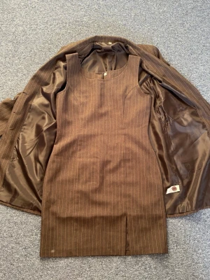 Mörkgrå ärmlös guldrandig kortklänning och tillhörande matchande kavaj - Snygg mörkgrå kortklänning med tunna, guldiga vertikala ränder och ärmlös design. Klänningen har en figurnära passform och rundad halsringning. Tillhörande säljs även den tillhörande, matchande kavajen i samma stil. Perfekt för dig som gillar en stilren och klassisk look. Oanvända och utan defekter, fläckarna är endast damm då de inte kommit till användning. 