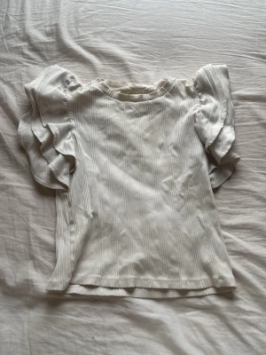 Vit topp med volangärmar - Supersöt vit topp från Gina Tricot med ribbad struktur och stora volangärmar som ger en extra stylish look. Perfekt för dig som vill ha något unikt och trendigt i garderoben. Rund halsringning och mjukt material som känns skönt mot huden.