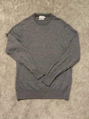 Grå tröja i merinoull från Calvin Klein - Stilren grå tröja från Calvin Klein med rund halsringning och diskret CK-logga broderad på bröstet. Tillverkad i tunt, mjukt material som ger en clean look. Perfekt till jeans eller chinos för en enkel men snygg outfit. Merinoull tror jag.