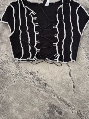 Svart croppad topp med snörning H&M - Trendig svart croppad topp från H&M med vita vågiga ränder och snörning framtill. Toppen har korta ärmar och ribbad struktur som ger en cool look. Perfekt för dig som gillar edgy detaljer och vill sticka ut.