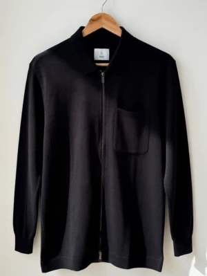 Svart kofta med dragkedja från Bläck storlek S - Stilren svart kofta från Bläck i storlek S. Koftan har en hel dragkedja framtill, klassisk krage och en bröstficka. Perfekt för dig som gillar minimalistisk design och vill ha något enkelt att slänga på sig. Materialet känns mjukt och bekvämt.