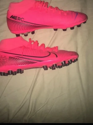 Nike Mercurial rosa fotbollsskor - Säljer ett par Nike Mercurial fotbollsskor i en riktigt snygg rosa färg med svarta detaljer och logga. Skorna har hög krage för extra stöd runt ankeln och är tillverkade i ett lätt syntetmaterial som ger bra bollkontroll. Perfekta för spel på konstgräs.