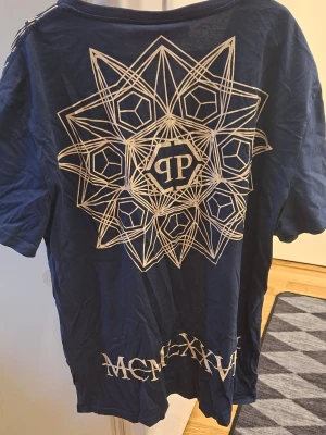 Blå Philipp Plein t-shirt med dödskallar - Snygg blå t-shirt från Philipp Plein med v-ringning och korta ärmar. Framsidan har två stora dödskalletryck, färgglada detaljer och logotyp. Baksidan har ett stort geometriskt mönster och romerska siffror längst ner. Perfekt för dig som gillar statement-plagg.